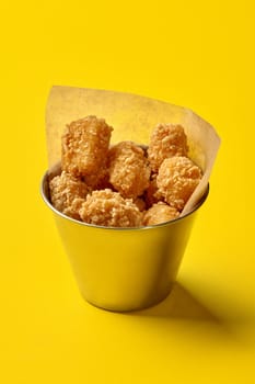 Metal bucket lined with parchment paper, filled with crispy chicken nuggets presented on vibrant yellow background. Concept of popular fast food snack