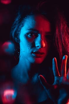 Sensual portrait of a beautiful attractive girl looking through the glass with raindrops with red blue lighting