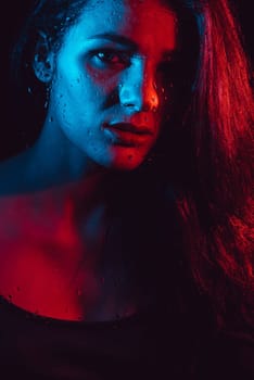 Sensual portrait of beautiful attractive brunette girl behind glass with raindrops with colored red blue neon lighting