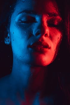 Sensual beauty fashion portrait of sexy attractive brunette girl through glass with raindrops and red blue neon lighting