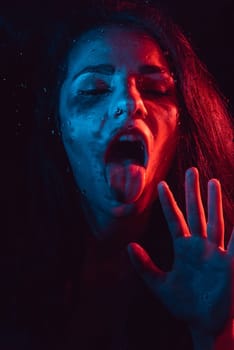 Sensual sad portrait of a girl with her tongue sticking out through the glass with raindrops and red blue lighting