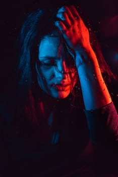 Sensual portrait of beautiful attractive brunette girl behind glass with raindrops with colored red blue neon lighting