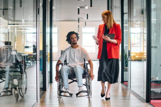 A business leader with her colleague, an African-American businessman who is a disabled person, pass by their colleagues who work in modern offices.