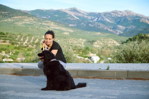 Smiling young woman sitting with her dog in the mountains, enjoying a happy time with her pedigree black cocker spaniel pet, dreamily looking aside into the distance. People and playing pets