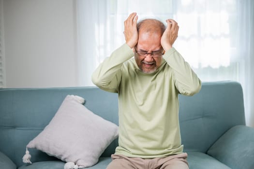 Headache. Sad Asian senior man sitting on sofa feeling hurt and lonely, elderly holds head with hand suffering from migraine headache, Old age health problems, healthcare