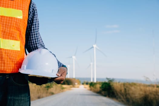 Engineer with safety helmet stands at wind turbine field. Represents renewable success innovation in combatting global warming. Displays industry leadership safety dedication.