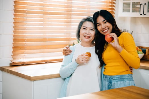 In the house, an old mother and her daughter, a young woman, stand together in the kitchen. They are holding an apple, showcasing the joy of learning and teaching, family, and togetherness.