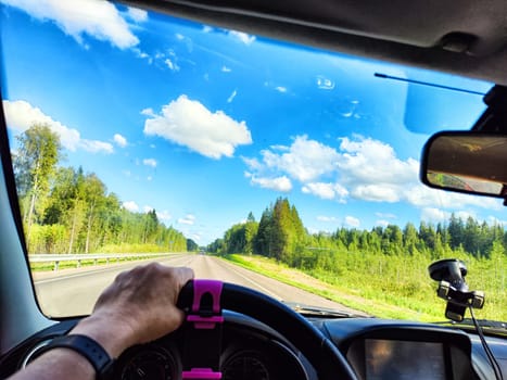 Car salon, steering wheel, hand of woman, view on nature landscape. View from seat of driver on Road, forest, blue sky, white clouds at sunny day. Concept of single trip of female traveller