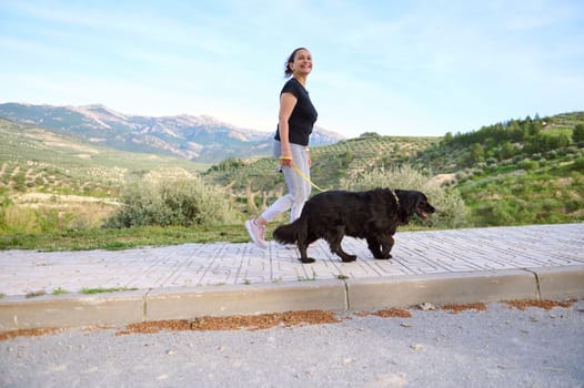 Full length shot of a woman in active wear, walking her dog on the mountains nature. Domestic animals, pets and people concept