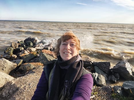 Happy cheerful middle aged woman in scarf taking selfie on nature outdoors and sun with water and waves of sea on the background