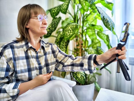 Smiling woman in glasses using a smartphone on a tripod for a video chat. A middle-aged woman posing and taking a selfie