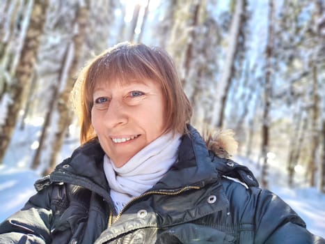 A cheerful middle aged woman in a winter coat taking selfie on nature outdoors in sunny day with blue sky