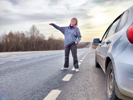 Girl Resting on Road Trip at Dusk by Her Car. A traveler takes a break, leaning on her parked car on an open road, with the sky hinting at dusk