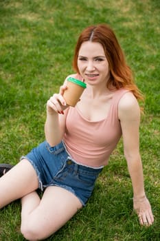 A beautiful young woman with braces on her teeth drinks from a cardboard cup while sitting on the grass. Vertical photo