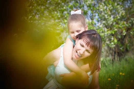 Happy mother and daughter enjoying rest, playing and fun on nature on a green lawn with dandelions and blooming apple tree on background. Woman and girl resting outdoors in summer and spring day