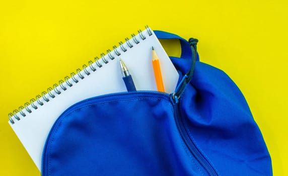 School backpack, pencil, pen and notepad on a yellow background. School blue backpack with notepad, pencil and pen inside on a yellow background.