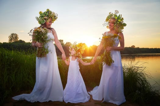 Ivan Kupala Celebration. Three Girls With Floral Wreaths by the River at Sunset. Family clad in white dresses celebrate Ivan Kupala by river at dusk.