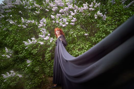 Elegant senior mature Woman in Black Dress looking like a witch by Blooming Lilac Bush at Dusk. Woman with red hair stands poised among lilac blooms