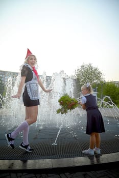 Young and adult schoolgirl on September with flowers having fun near water of fontain. Generations of schoolchildren, pioneer of USSR and October girl in modern uniform of Russia. Mom and daughter