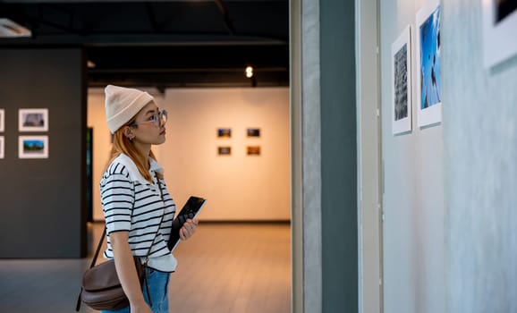 Asian woman holding tablet at art gallery collection in front framed paintings pictures on white wall, Young person at photo frame hold digital book leaning against at show exhibition artwork gallery