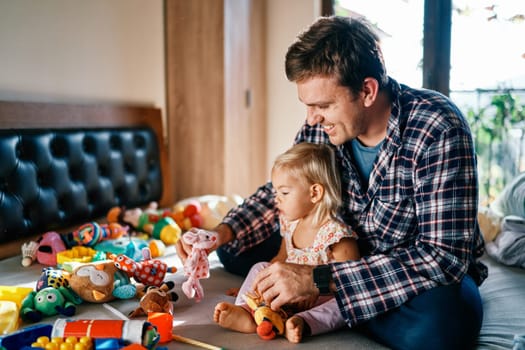 Dad sits behind a little girl and plays soft toys with her. High quality photo