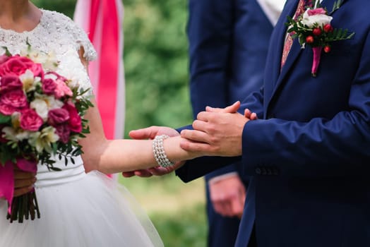 groom in a blue suit holds the bride's hand in a white dress at the wedding ceremony