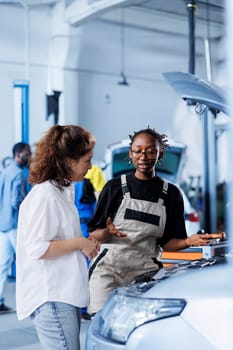 Trained mechanic helping client with car maintenance in auto repair shop after her car shut down unexpectedly. Employee in garage mending customer damaged vehicle fuel tank