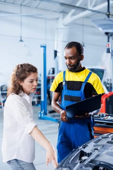 Smiling serviceman in car service uses tablet to calculate invoice after fixing broken automobile radiator. Specialist uses device to inform customer of final costs after mending vehicle