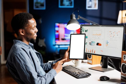 Young person working on tablet with blank display, sitting at his workstation before evaluating accounting statistics on pc. African american man holding device with white screen template at home.
