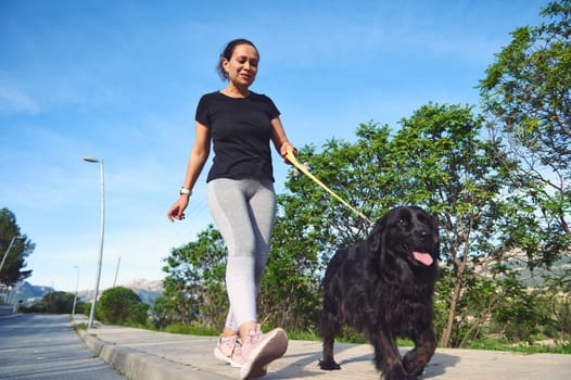 View from the bottom of a multi ethnic happy young woman in sports clothes, walking her dog on leash and enjoying morning run with her pet in the mountains nature. People and playing pets concept