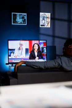 Curious spectator reads news coverage titles on television, sits on couch and listens to journalist lay out recent global events. Young person watching daily news headlines, tv content.