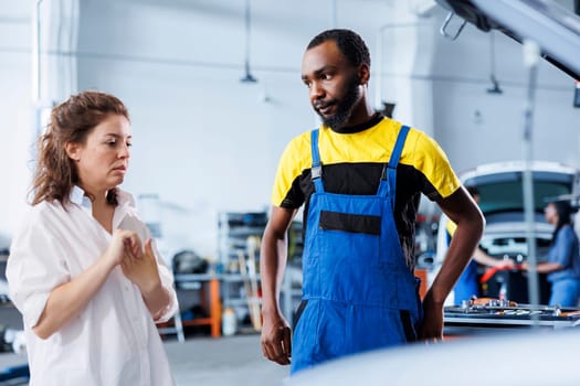 BIPOC mechanic helping client with car maintenance in auto repair shop. Employee in garage facility looking over automobile parts with woman, mending her vehicle motor during inspection