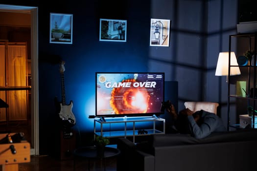 Player is dissatisfied after failing to win shooter tournament while engaging in web based multiplayer battle at night. Unhappy adult playing first person online action games on home television.