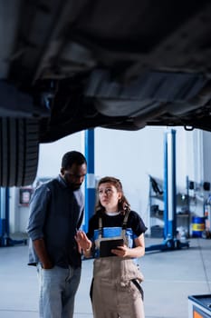 Knowledgeable mechanic in garage using laptop to follow checklist while doing maintenance on car, talking with client. Specialist in auto repair shop does checkup on vehicle tires next to customer