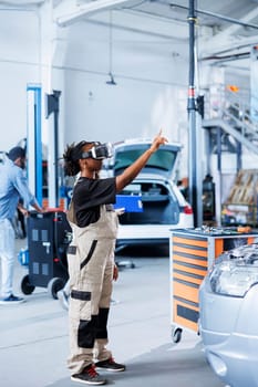 Licensed engineer in auto repair shop using virutal reality goggles to visualize car components in order to fix them. African american woman using VR technology while working on damaged vehicle engine