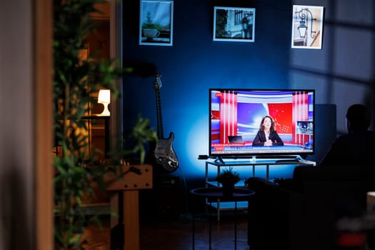 Relaxed person watching breaking news on television at night, chilling on sofa in living room and watching tv program. Young adult listening to media information on global network.