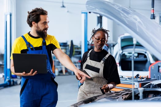 Technician servicing broken vehicle, checking for faulty brakes under manager supervision. Smiling repairmen in auto repair shop working together on fixing automobile, discussing best options