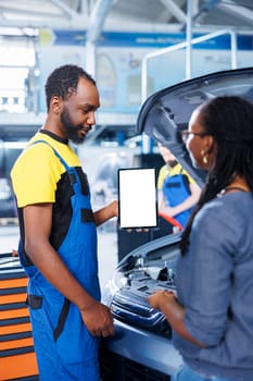 Smiling engineer in car service uses mockup tablet to order new compressor belt for damaged vehicle. Employee showing client needed components replacement on isolated screen device
