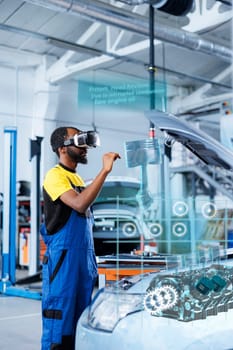 BIPOC mechanic in repair shop using augmented reality holograms to check car performance parameters during maintenance. Qualified garage employee using futuristic vr headset to examine damaged vehicle