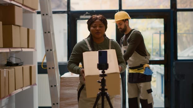 Warehouse supervisor pressing play on cellphone placed on tripod to start recording training video for interns. Quality assurance manager filming tutorial showing trainees how to arrange parcels