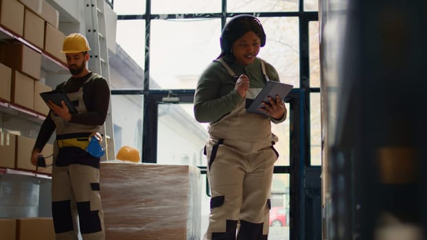 Relaxed african american warehouse picker listening music while checking labels on cardboard boxes. Smiling worker wearing heaphones and bopping head while walking through storage area aisles