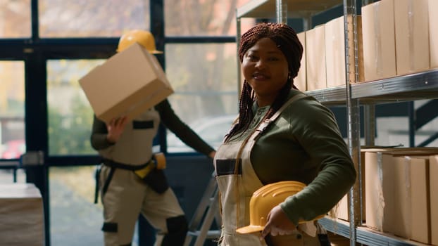 Portrait of cheerful african american warehouse sorter in protective uniform dealing with packaged stock in cardboard boxes ready to be shipped, happy to work in professional distribution center
