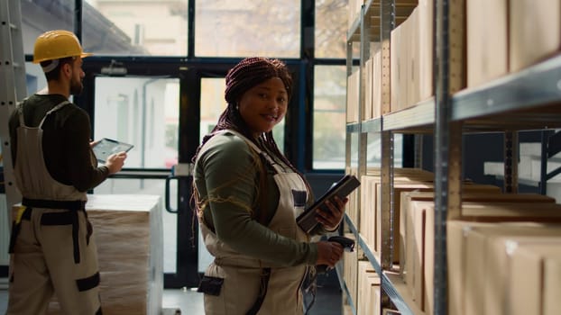 Dolly in portrait shot of cheerful african american warehouse employee scanning cardboard boxes on shelves, updating products labels info before sending parcels to be shipped