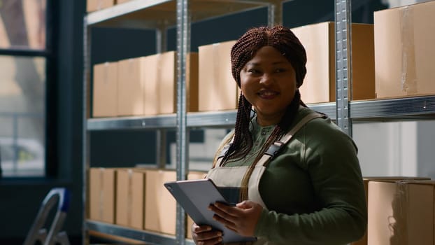 Portrait of joyful warehouse chief supply chain officer holding tablet, checking packaged goods in cardboard boxes, ensuring errorless operations in professional storage room