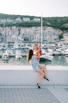 Mom picks up a little laughing girl in her arms while sitting on the curb of a marina with moored yachts. High quality photo
