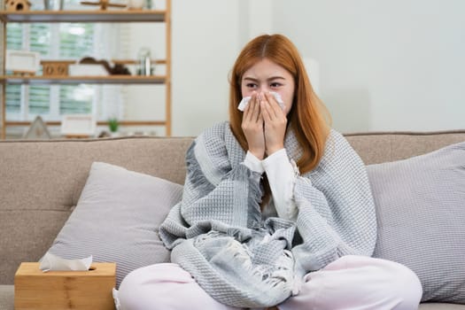 young woman sitting on sofa covered with blanket freezing blowing running nose got fever caught cold sneezing in tissue.