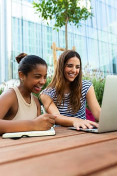 Two happy female college multiracial friends studying together in campus using laptop. Vertical image. Education and technology concept.