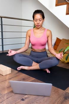 Young Asian woman meditating at home with online video meditation lesson using laptop. Vertical image. Healthy lifestyle concept.