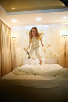 Cute little girl jumping on white bed in sleeping room or in hotel in evening