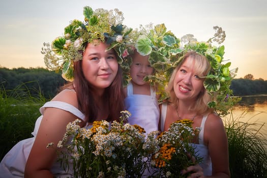 Ivan Kupala Celebration. Three Girls With Floral Wreaths by the River at Sunset. Family clad in white dresses celebrate Ivan Kupala by river at dusk.